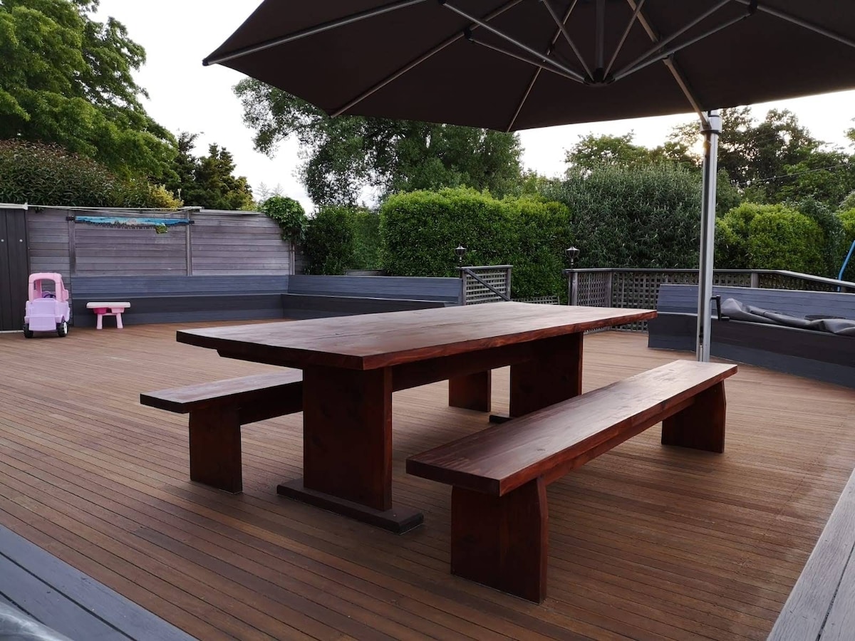 A large wooden table with matching benches is situated on a spacious deck, surrounded by lush greenery. An umbrella provides shade, while wooden flooring enhances the outdoor setting. A pink toy kitchen is visible in the background, adding a playful touch to the area.