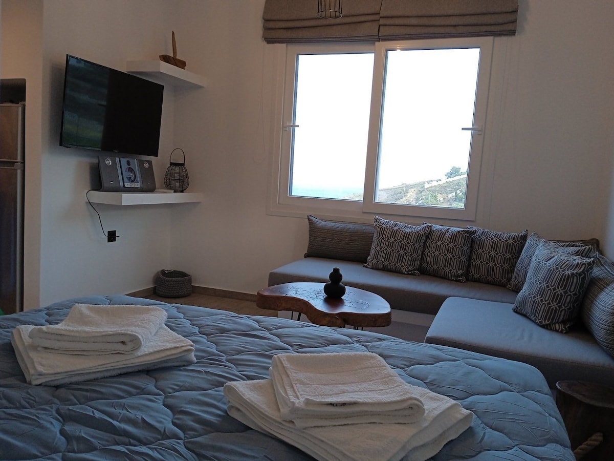 A well-appointed room features a spacious bed covered with neatly folded towels. A cozy seating area is situated next to large windows, offering views of the surrounding landscape. A flat-screen television rests on a minimalist shelf, contributing to the room's modern design.