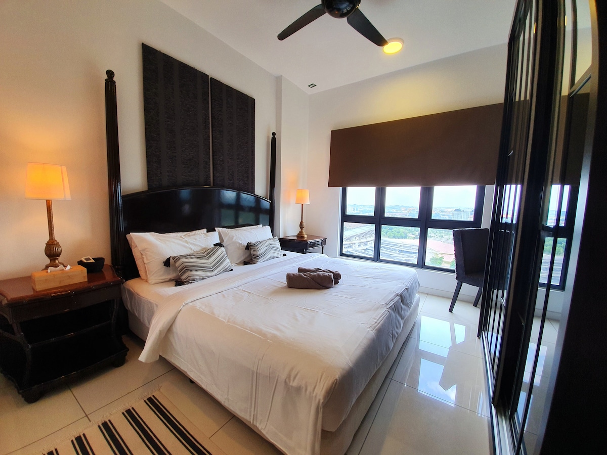 A well-appointed bedroom is featured, showcasing a spacious bed with striped pillows and a soft blanket. Natural light floods the room through large windows, which overlook scenic views. Additional furnishings include bedside tables and a seating area with a chair, complemented by stylish wall decor.