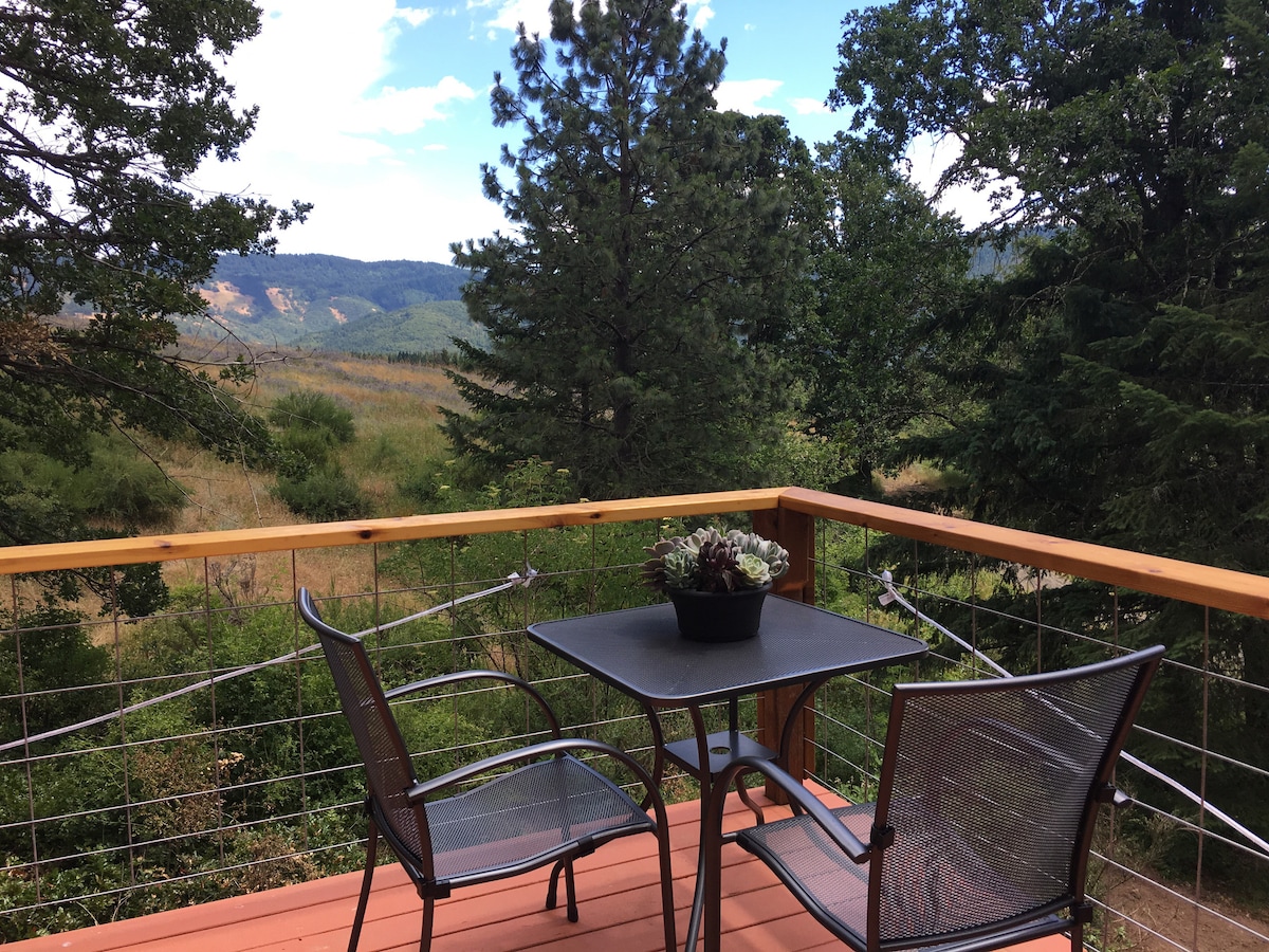 A small outdoor deck features a round table surrounded by two metal chairs. A potted succulent sits center on the table. The scenic view includes rolling hills, dense trees, and a clear blue sky with scattered clouds, creating a peaceful atmosphere.