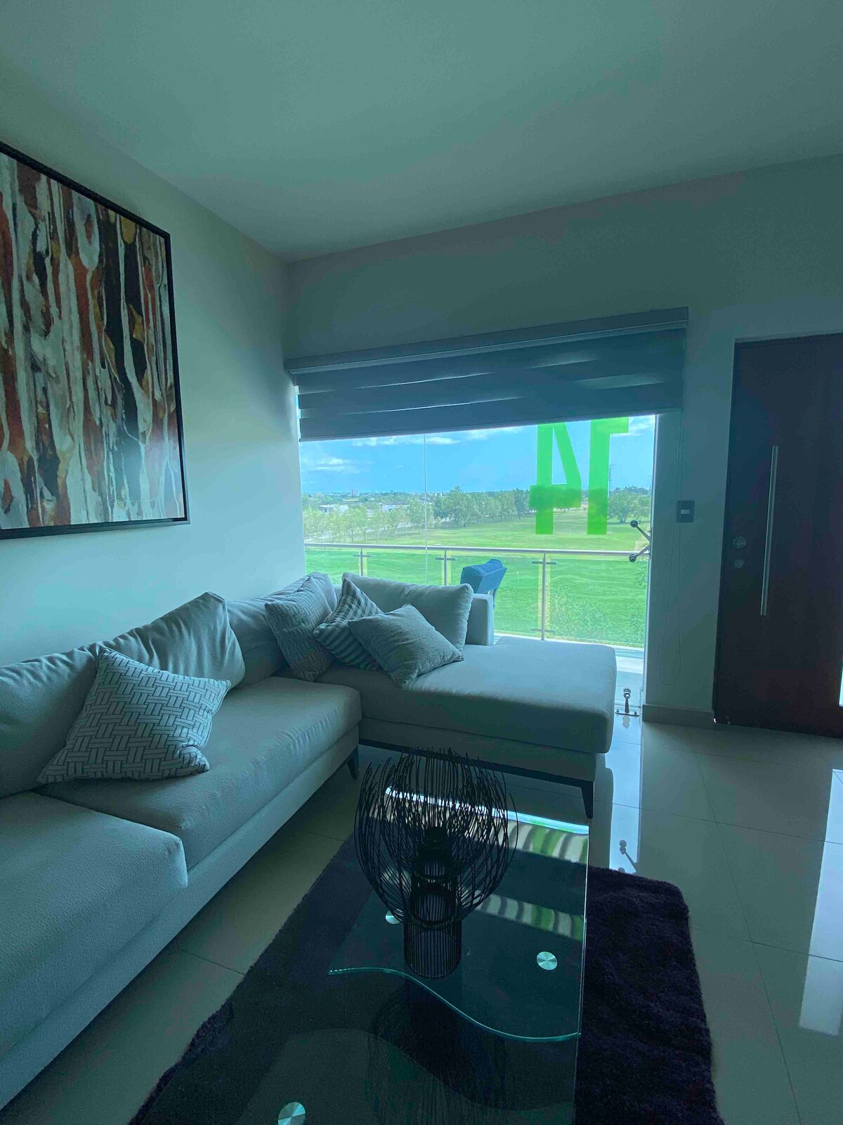 A modern living area features a light grey sectional sofa adorned with patterned cushions. A glass coffee table sits in front of it, while large windows provide views of the surrounding greenery and open landscape. Natural light enters through partially drawn shades.