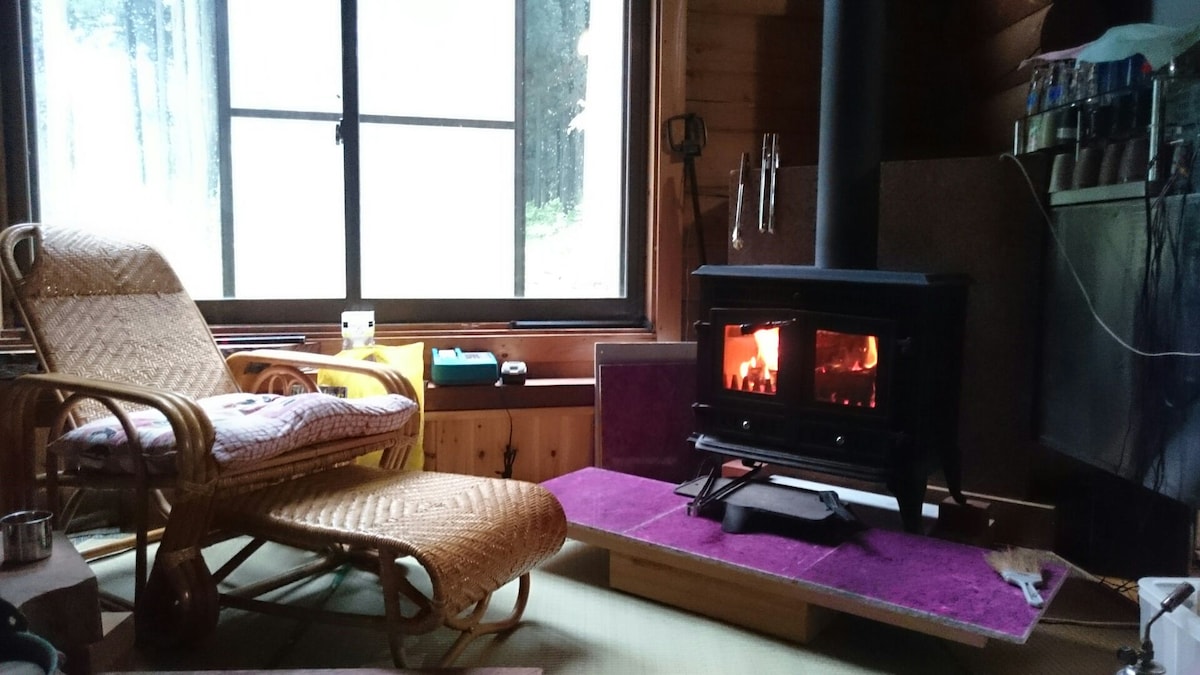 A comfortable seating area is highlighted by a wooden chair with soft cushions, positioned near a stove with a glowing flame. Large windows provide a view of the surrounding nature, while shelves display various items, enhancing the cozy environment.