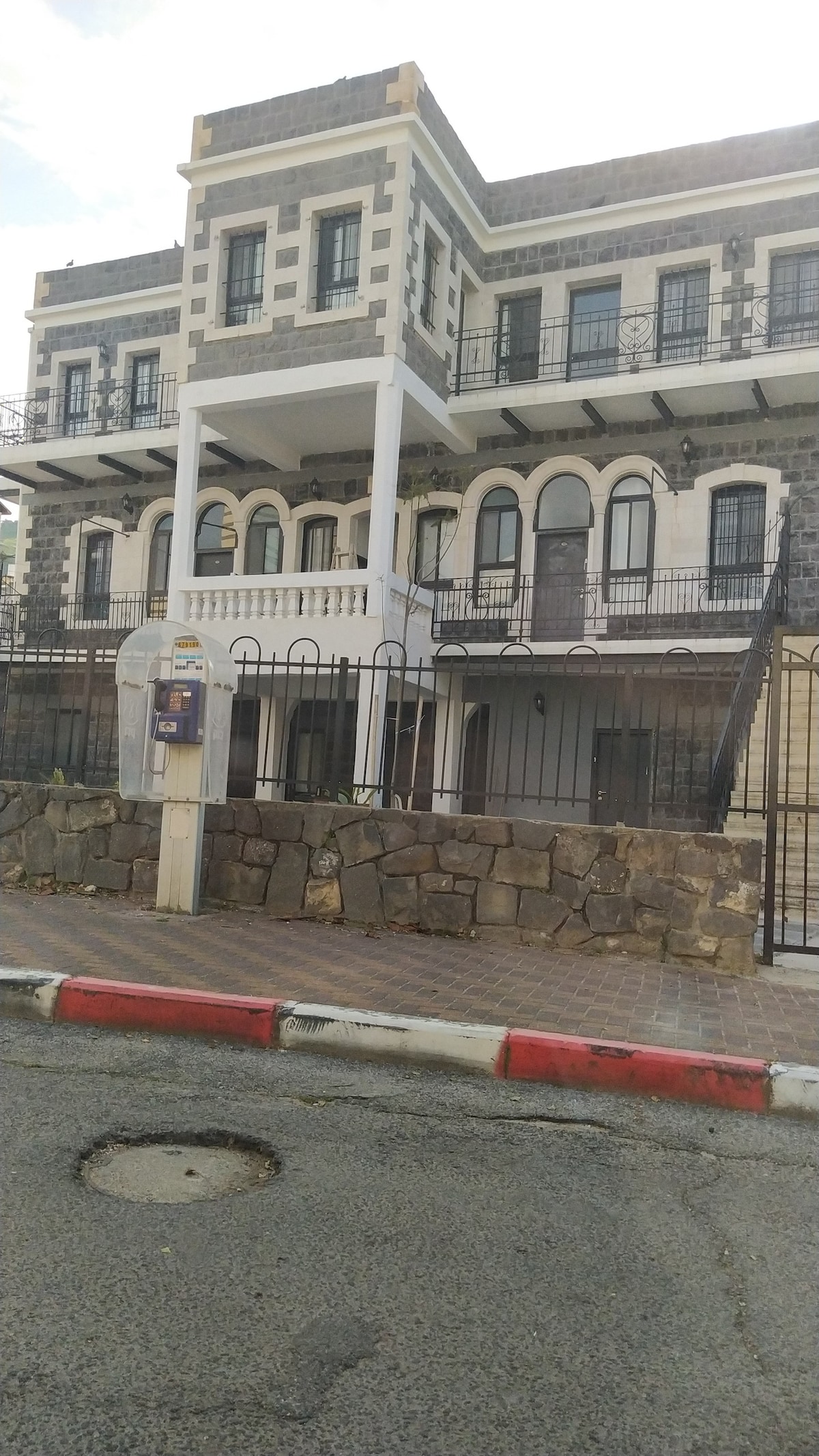 The exterior of a historic stone building is showcased, featuring a charming façade with intricate architectural details. A spacious balcony is visible on the second level, framed by ornate railings. Surrounding greenery and a paved pathway contribute to the inviting atmosphere of the property.