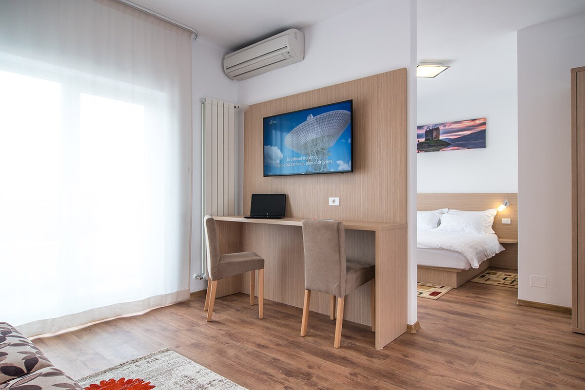 A bright lounge area features a sleek desk and two upholstered chairs, with a 50" Smart TV mounted on a wooden wall. Natural light filters in through sheer curtains, illuminating the space that leads to a comfortable bedroom area beyond.