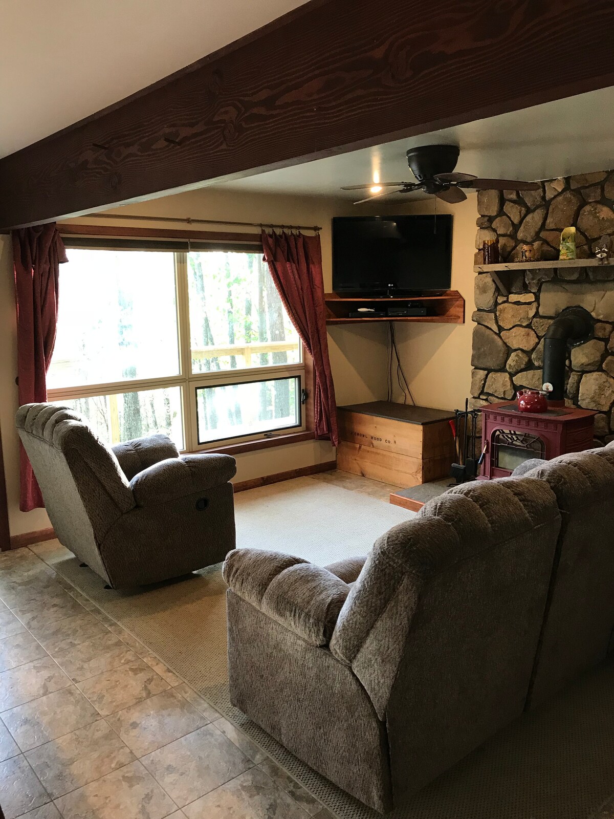 A cozy living area features two comfortable recliners and a plush sofa facing a stone accent wall with a small wood stove. A flat-screen TV is mounted above a wooden shelf, while large windows framed by burgundy curtains allow natural light to fill the space.