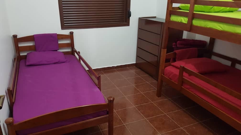 The room contains a twin bed with a purple mattress and a wooden frame, alongside a bunk bed with green bedding on the upper and red bedding on the lower level. A brown dresser stands against the wall, and a window with horizontal blinds provides natural light.