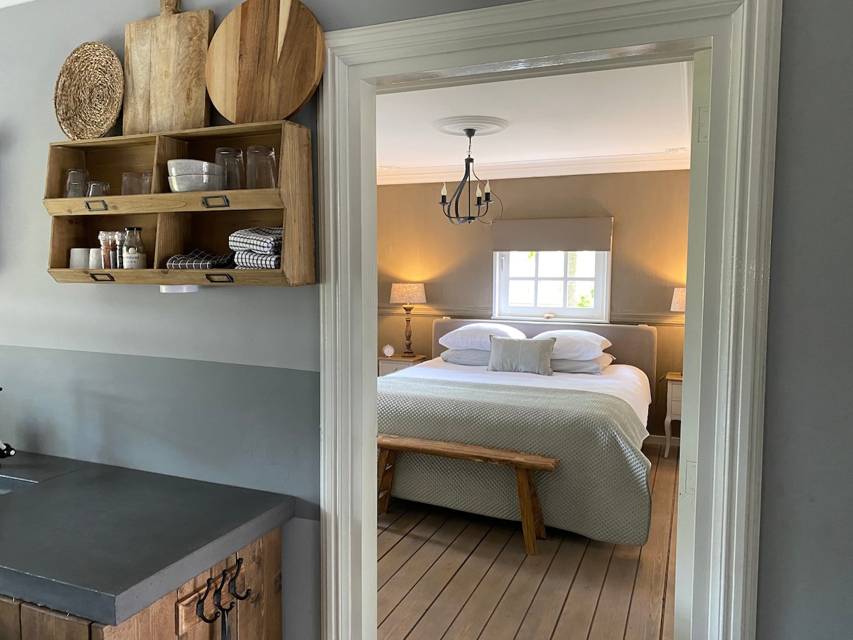A doorway reveals a cozy bedroom featuring a spacious bed with soft white pillows. Warm lighting from bedside lamps enhances the natural wood tones, while wooden flooring complements the inviting space. A shelf holds various kitchenware, showcasing the connection between the kitchen and the sleeping area.