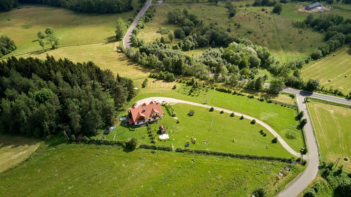 An aerial view of the property reveals a spacious green area surrounded by trees. The house with a red roof is situated centrally, with pathways leading to various outdoor spaces and play areas. The landscape features open fields and winding roads.