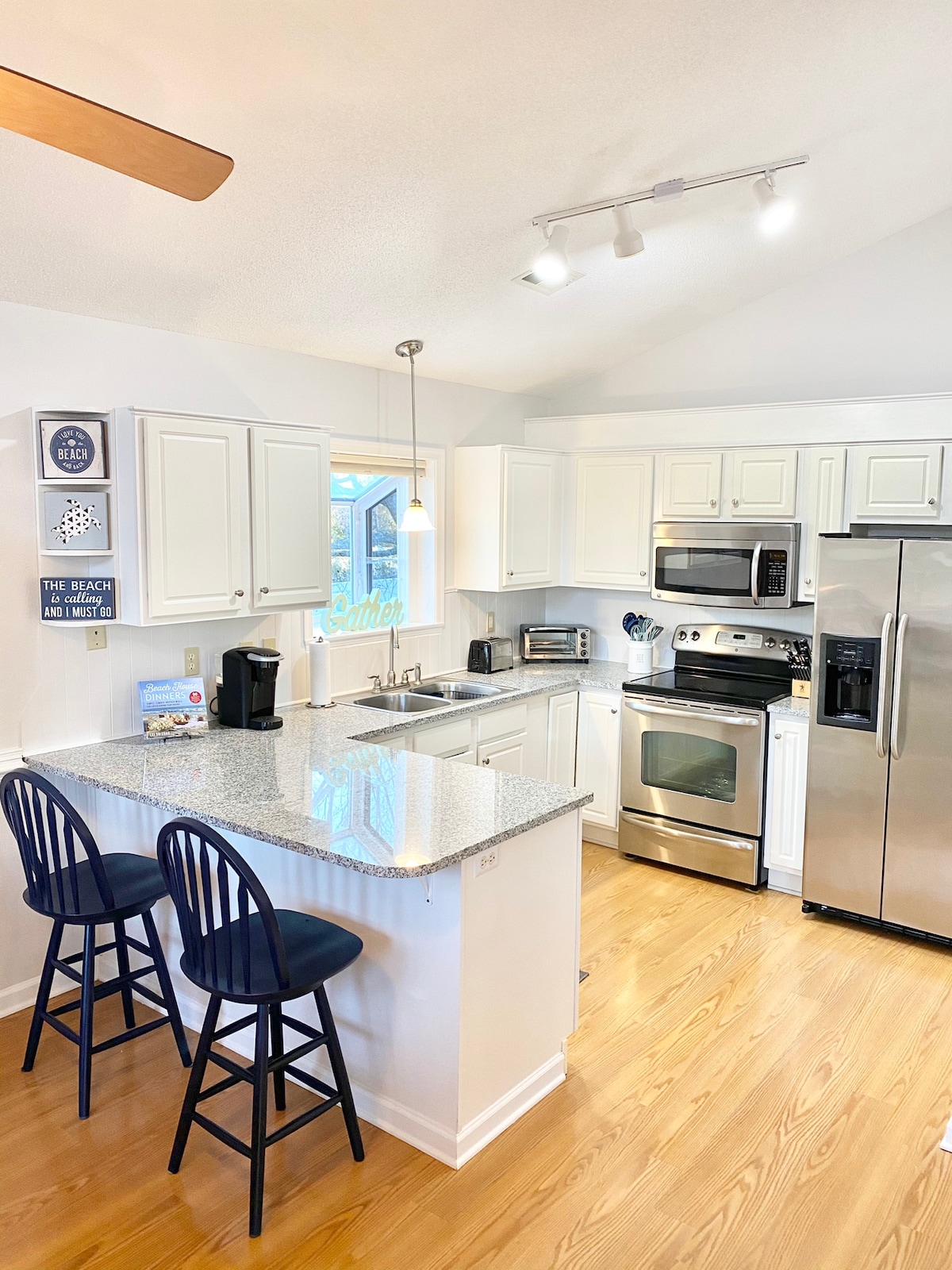 The kitchen features contemporary white cabinetry and stainless steel appliances. A central island with two dark barstools provides a casual seating area. Natural light fills the space through a nearby window, enhancing the clean and functional design.
