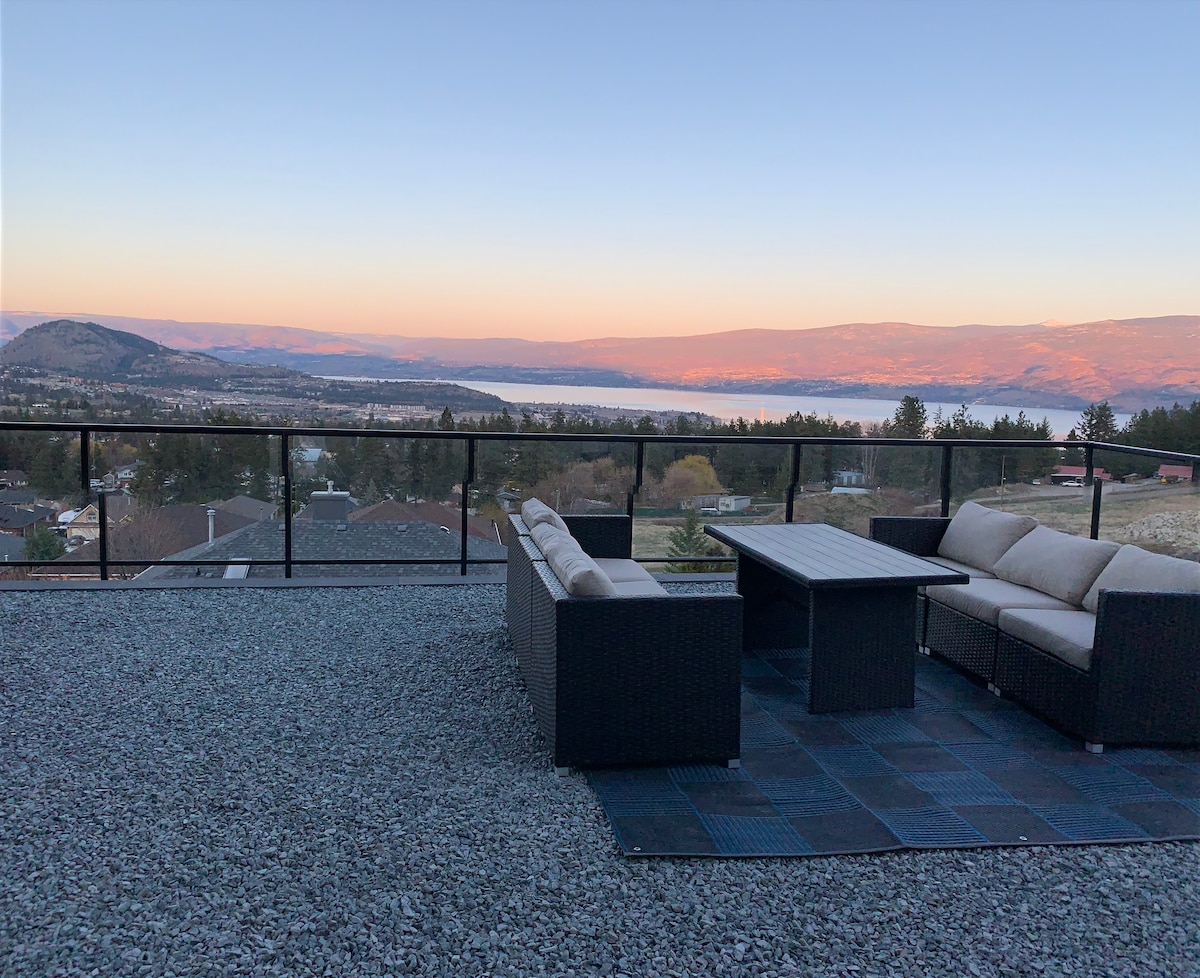 An outdoor seating area is presented on a private patio, featuring a dark woven sectional sofa and a matching coffee table. The space overlooks scenic views of Okanagan Lake and rolling hills during sunset, with a gravel surface adding texture beneath.