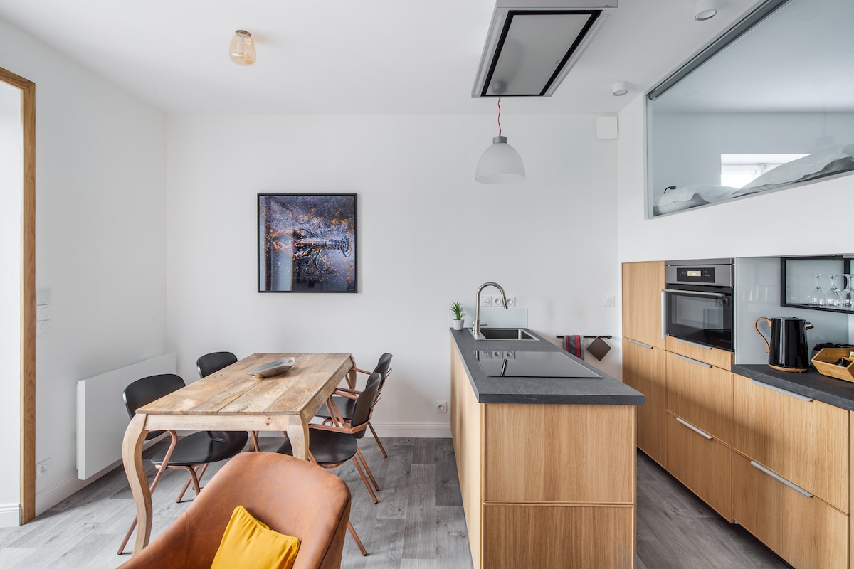 A modern kitchen and dining area showcase a mix of wooden textures and sleek surfaces. A large wooden dining table is complemented by six black chairs. The kitchen features an integrated oven, countertop appliances, and open shelving, creating a functional and inviting space.