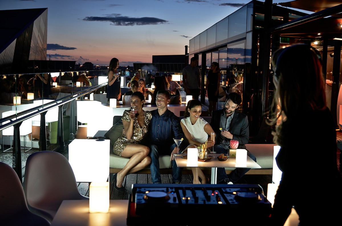 An outdoor rooftop setting is depicted with modern furniture and softly glowing table lamps. Guests are seated together enjoying drinks, while a DJ station is visible in the foreground. The sky transitions into twilight, with a subtle gradient of blue and orange tones.