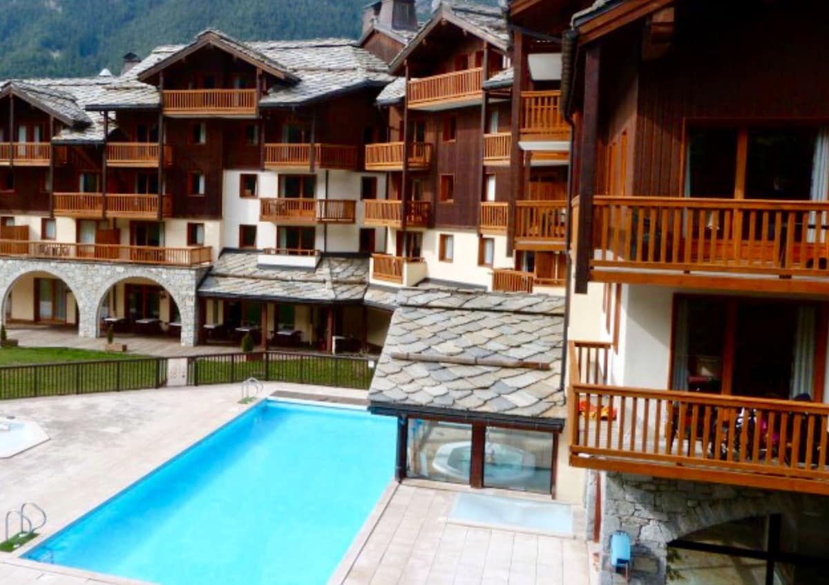 A communal outdoor pool is framed by a spacious deck and wooden balustrades. Surrounding the pool, multiple buildings display traditional alpine architecture, characterized by stone and wood materials. The scene is complemented by a well-maintained lawn and mountain views in the background.