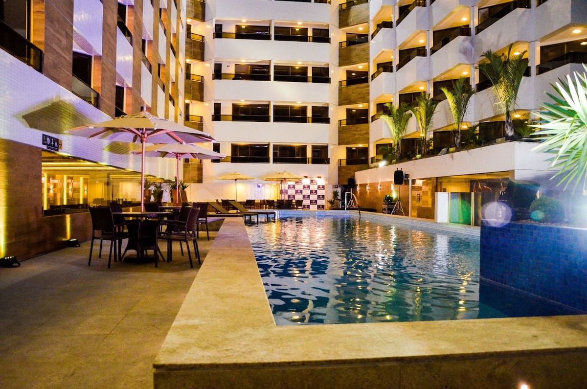 A well-lit pool area is framed by tall palm plants and outdoor seating. Umbrellas provide shaded spots near the pool, which reflects soft lighting from surrounding features. The structure has multiple levels, with balconies visible overlooking the pool.
