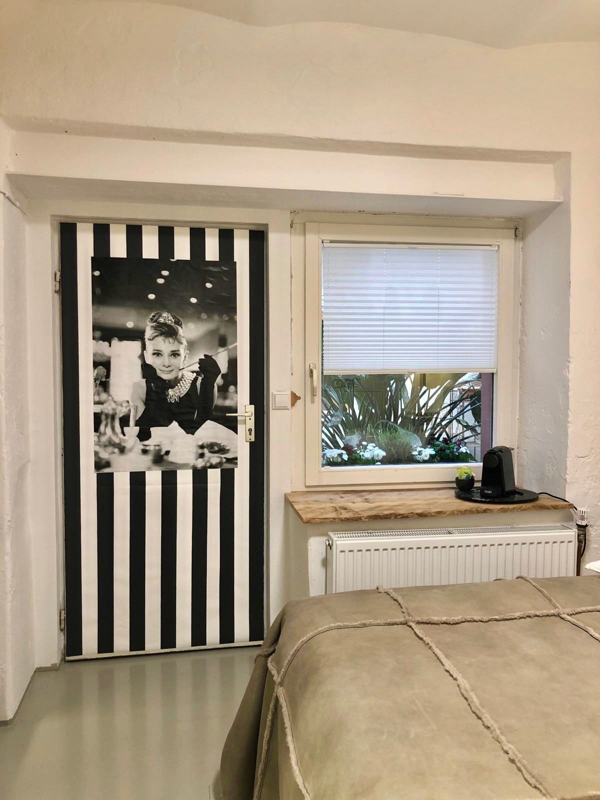A room is presented featuring a black and white striped door that contrasts with the white walls. A window is visible, decorated with light blinds, allowing natural light to enter. A small table with a coffee maker sits adjacent to the window, enhancing the minimalistic design.