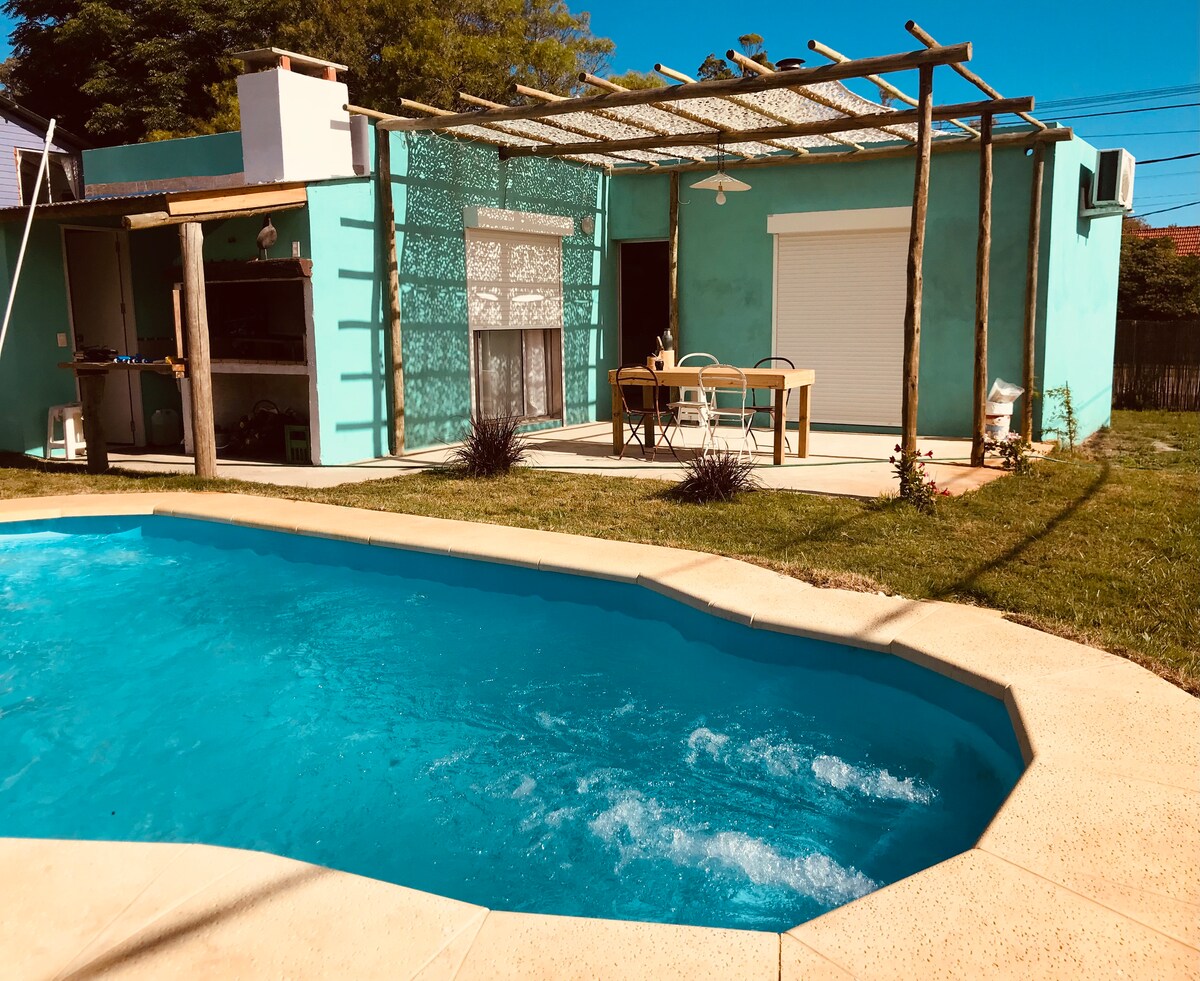 A refreshing swimming pool is framed by a spacious grassy area, with a covered outdoor dining set nearby. The house features a vibrant turquoise exterior and large windows, allowing natural light to illuminate the space. A barbecue area is visible in the background.