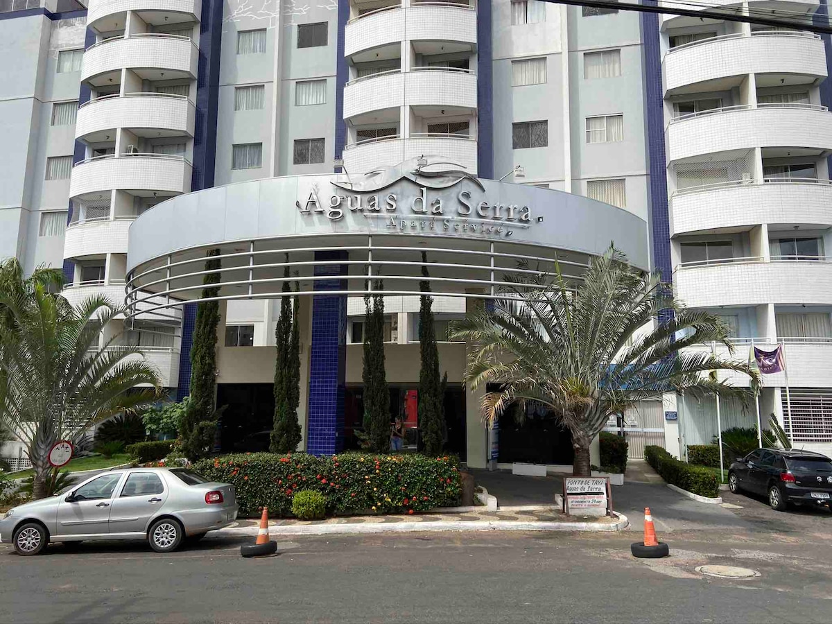 The entrance of the Aguas da Serra Apart Services is framed by neatly trimmed palm trees and landscaped greenery. The building features a modern design with multiple levels and a prominent sign at the top. A parked car and traffic cones are visible in the foreground.
