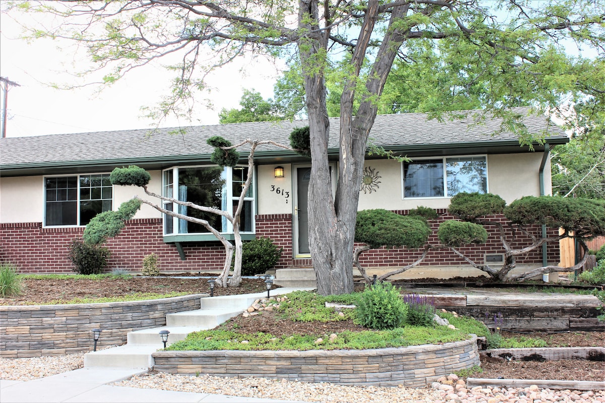 The exterior of a single-story home displays a blend of brick and stucco siding. Neatly landscaped areas feature low-growing shrubs and decorative stones. A large tree with a sculpted appearance provides shade, while a pathway leads to the entrance, enhanced by subtle outdoor lighting.