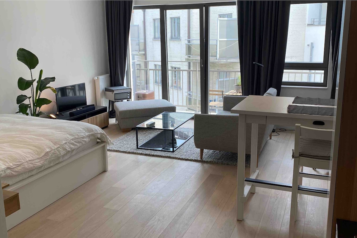 A spacious room is displayed featuring a large bed with a soft white comforter. Natural light fills the area through expansive glass doors, which open to a small balcony. A modern seating arrangement includes a sofa and a low table on a light rug.