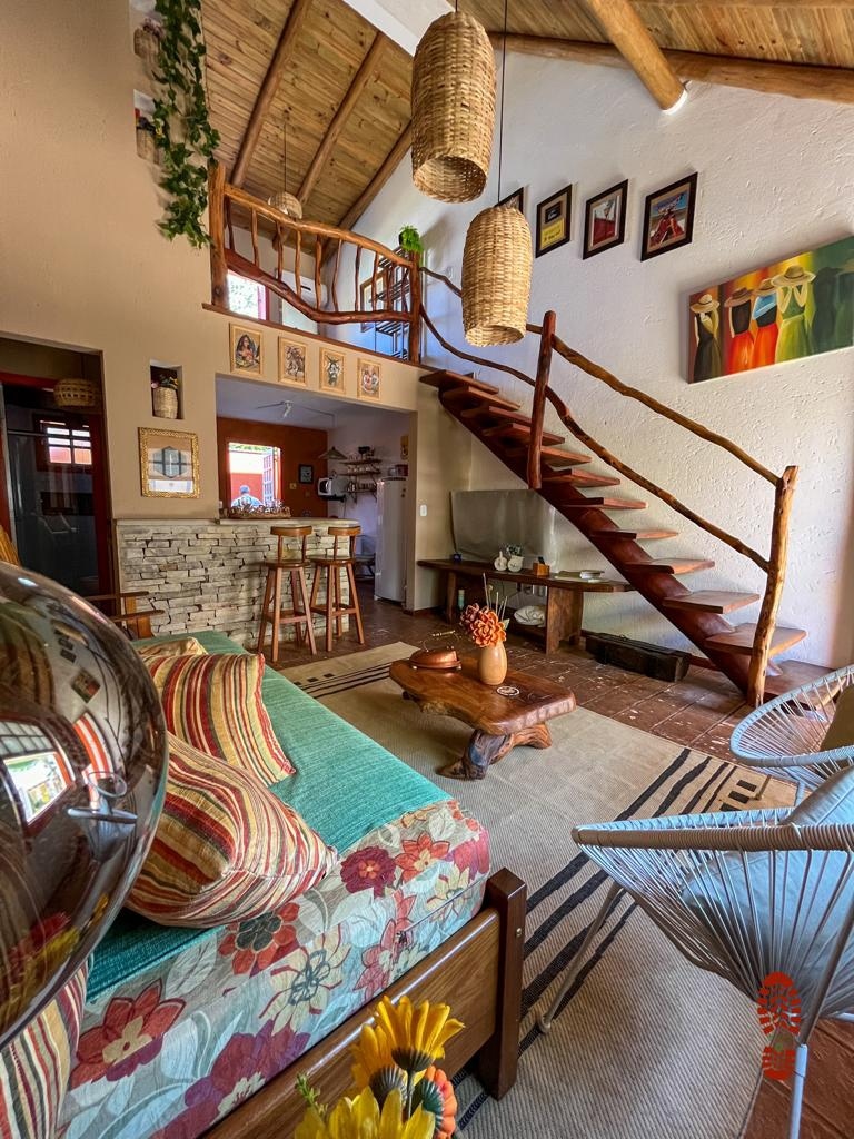 A spacious living area features a wooden staircase leading to the upper level, adorned with woven light fixtures. A cozy seating space is arranged with colorful cushions, while a rustic coffee table holds decorative items. Artworks line the walls, enhancing the homey feel.