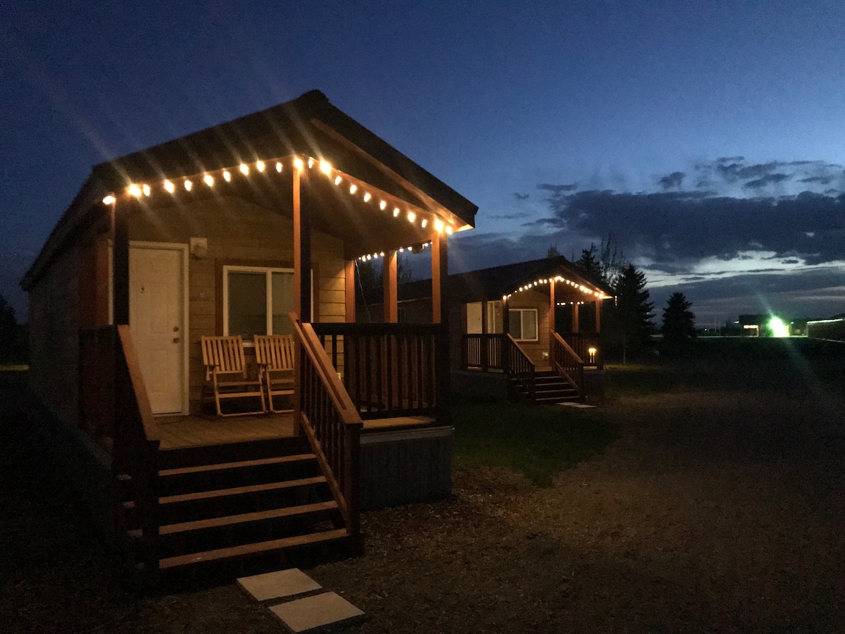 Two cabin structures are illuminated by string lights, creating a welcoming ambiance at dusk. Each cabin features a front porch with wooden rocking chairs and stairs leading up to the entrance. The surrounding area is open with a visible horizon and scattered clouds in the evening sky.