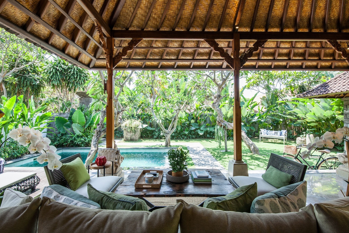 An open-air terrace features a wooden ceiling crafted with traditional Balinese design. Comfortable seating arrangements include plush sofas adorned with green accent pillows, while a central table holds decorative items. The lush tropical garden and private swimming pool are visible through the expansive outdoor space.