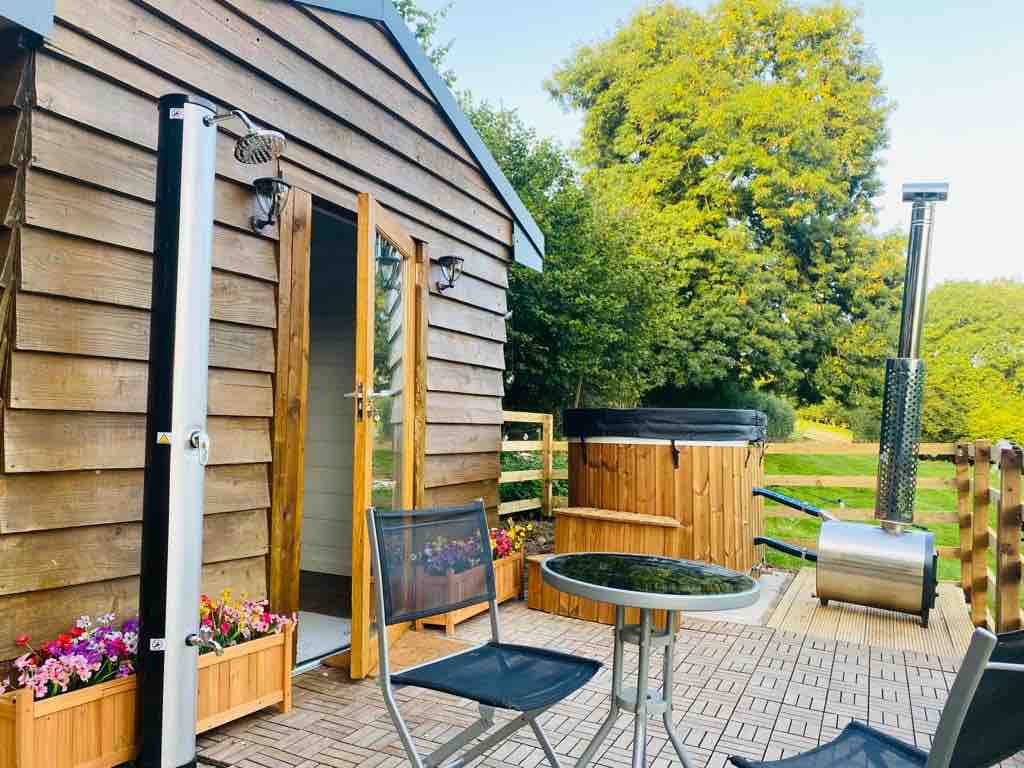A wooden cabin features a welcoming entrance door and is complemented by a cozy seating area with a small table and chairs. An outdoor log-fired bath is prominently positioned on the deck, surrounded by vibrant flowers and greenery, promoting a serene escape into nature.
