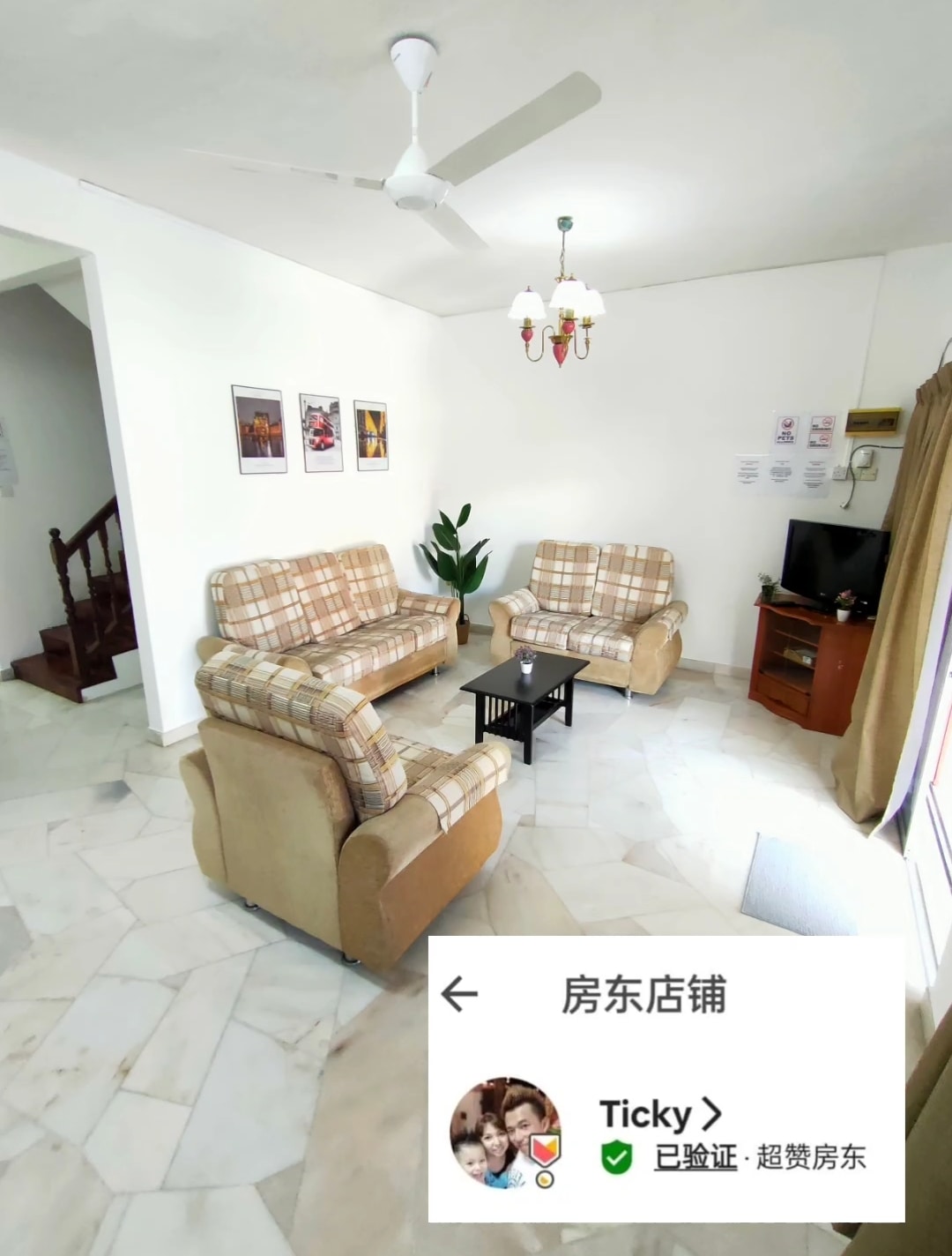 A spacious living area with a light-colored marble floor is furnished with two plaid sofas and a coffee table. A television sits beside a window dressed with neutral curtains. A leafy plant adds a touch of greenery, while photos are displayed on one wall.
