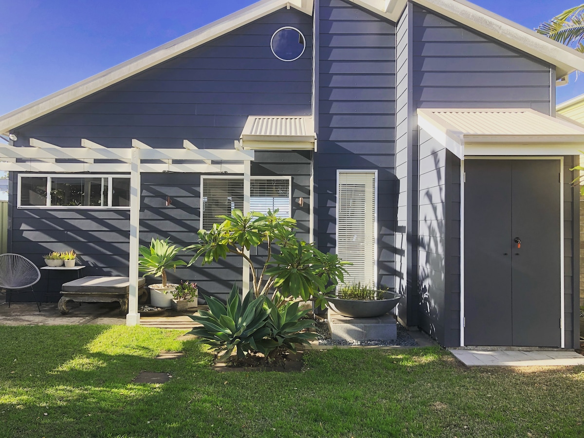 The exterior of the house showcases a modern blue façade with white trim. A pergola provides shade over a sitting area, surrounded by green plants. Windows allow natural light to filter in, while a gray door offers access to the interior.