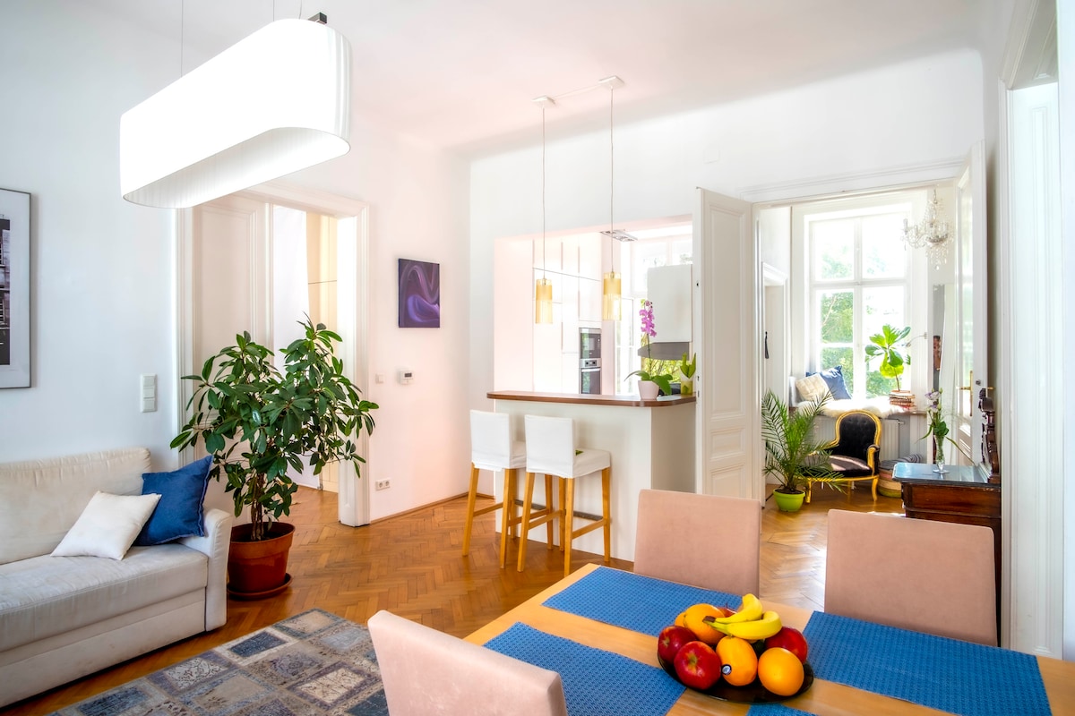 A spacious living area is illuminated by a modern light fixture. A dining table with bar stools is positioned next to a well-equipped kitchen. A sofa is placed nearby with a colorful blue pillow. Natural light filters through large windows, complemented by indoor plants.