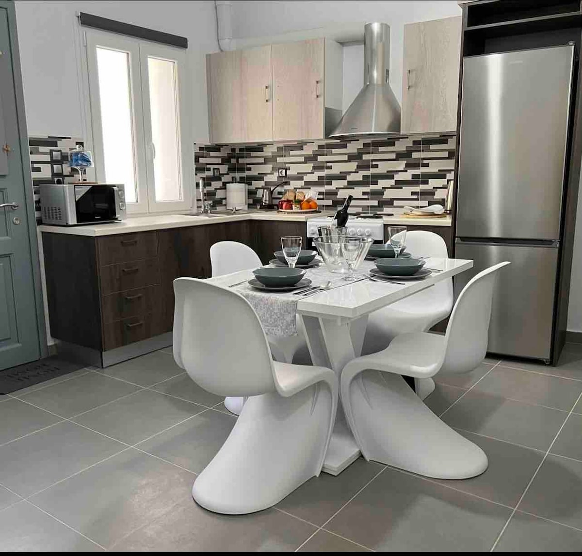 A modern kitchen is depicted, featuring sleek cabinetry and a geometric dining table surrounded by six white chairs. Kitchen appliances, including a stainless steel refrigerator and a stovetop, are present. The backsplash is decorated with a mosaic of gray and black tiles, enhancing the contemporary design.