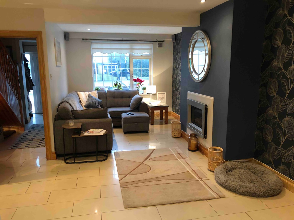 A comfortable living room is presented, featuring a gray sectional sofa with a matching ottoman. A circular mirror reflects natural light from a nearby window. Soft lighting enhances the atmosphere, and a rug lies on the tiled floor alongside decorative elements like a lantern and pet bed.