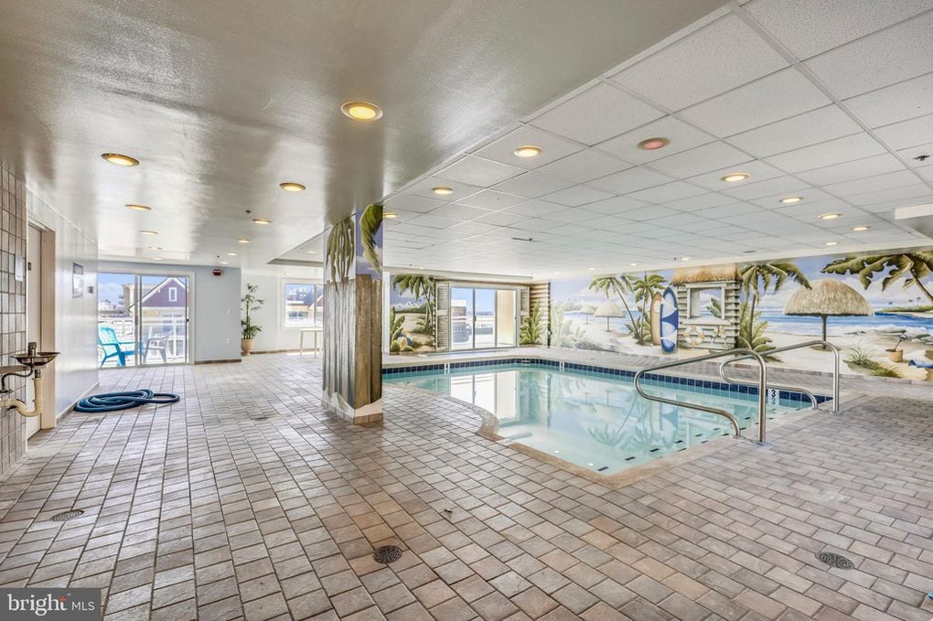 The indoor pool area is bright and spacious, featuring tiled flooring and large windows that offer views of the outdoors. A tropical mural adorns the walls, enhancing the beach-themed ambiance. A ladder provides access to the pool, surrounded by lounging space.