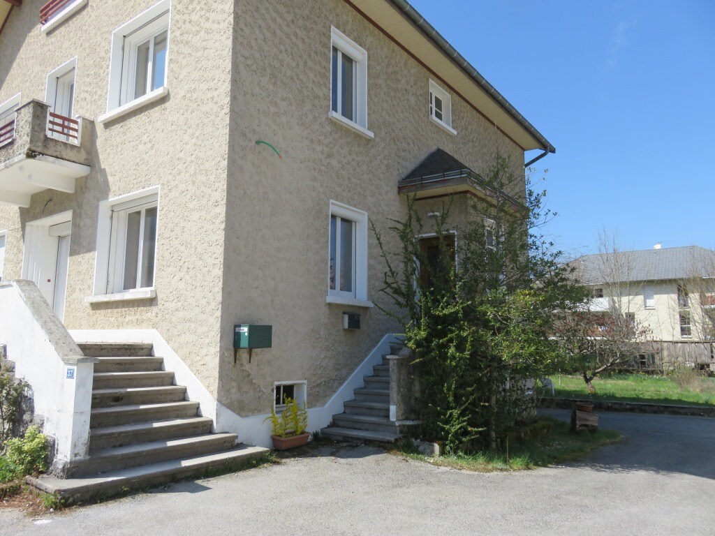 The exterior of a two-story building is visible, featuring light-colored walls and multiple windows. A set of concrete steps leads to the entrance, with a small shrub and planter near the base. The surrounding area is tranquil, showcasing greenery and a clear blue sky.