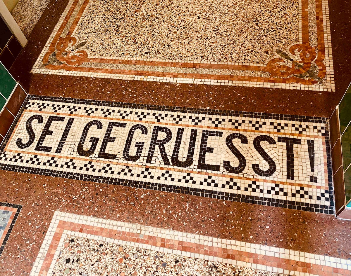 A mosaic floor welcomes visitors with the phrase 'SEI GEGRUESST!' prominently displayed. The intricate tile design features a blend of colors and patterns, showcasing the elegance of the entryway's historical architecture.