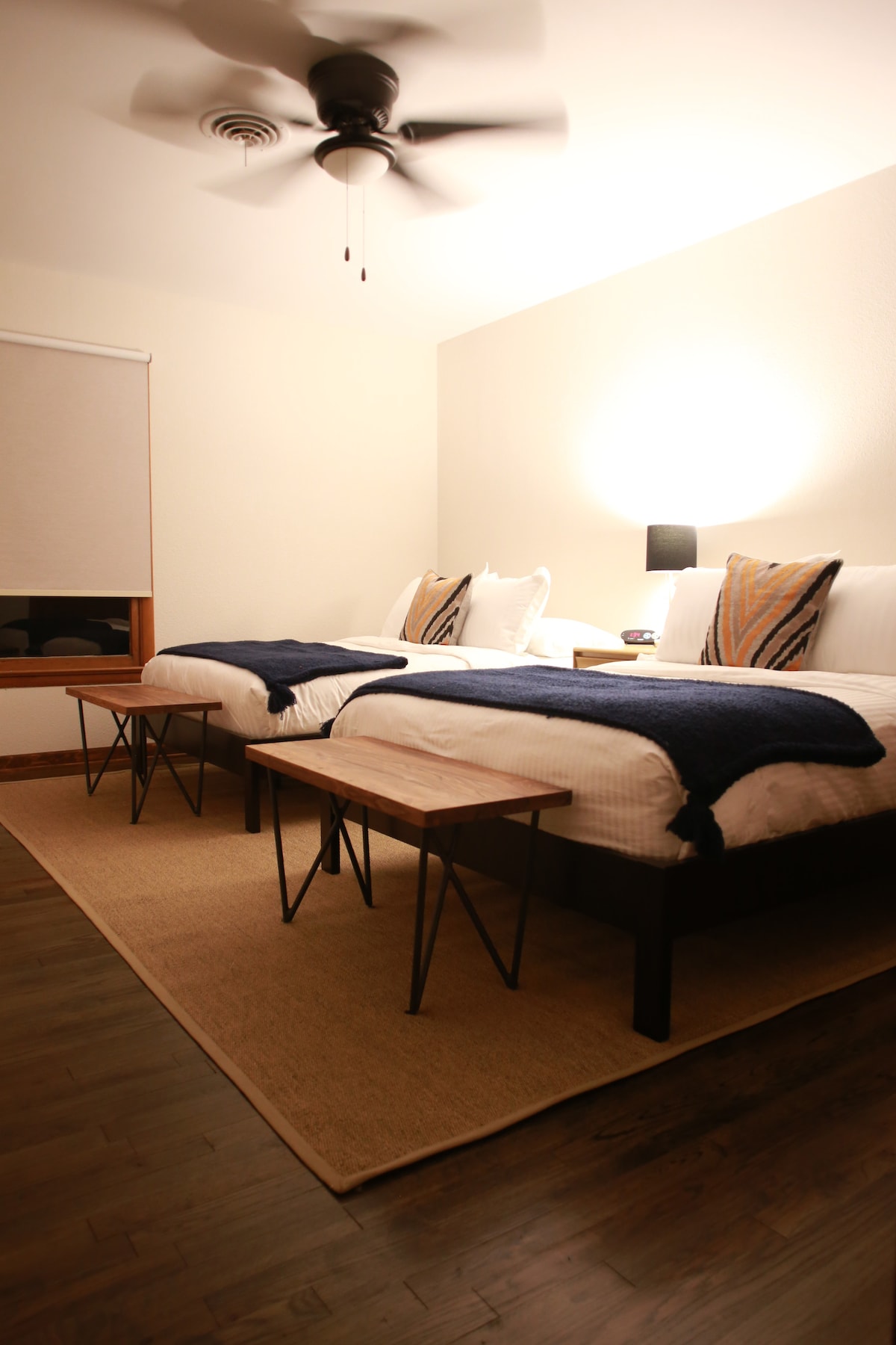 The room features two spacious beds with modern bedding and decorative pillows. A table is positioned between the beds, providing a functional space. Soft lighting is provided by a bedside lamp. Natural light filters through a window covered by a light shade.