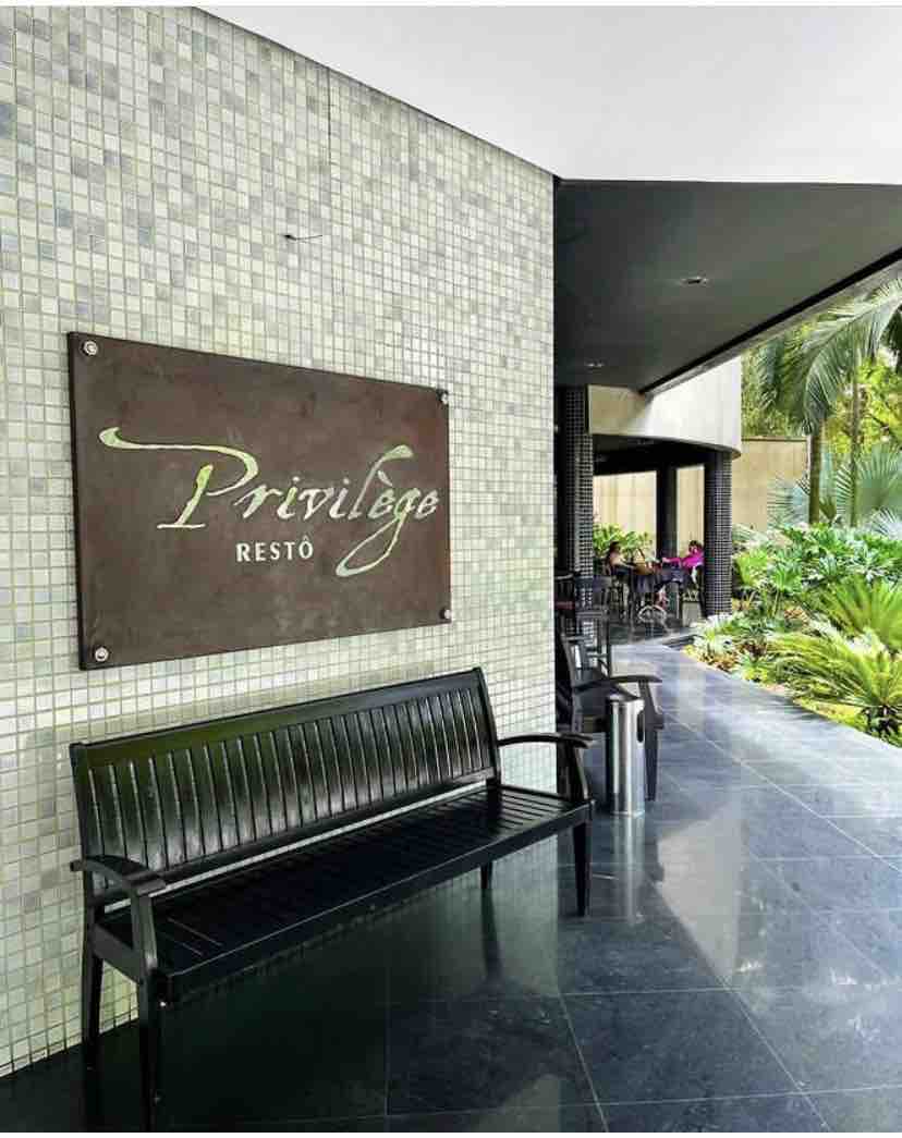 A modern entryway features a sleek black bench alongside a tiled wall with the sign 'Privilege Resto'. Lush greenery and tropical plants are visible, providing a refreshing ambiance. The polished floor reflects the natural light illuminating the space.