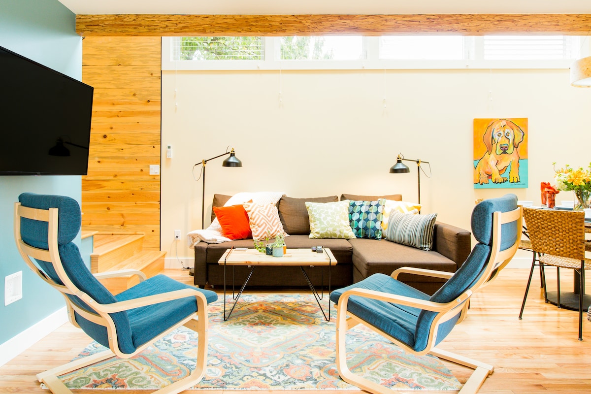 A cozy living area features a sectional sofa adorned with colorful cushions, paired with two blue armchairs. A large piece of artwork depicting a dog is displayed on the wall. Light floods the space through multiple skylights, creating a welcoming atmosphere.