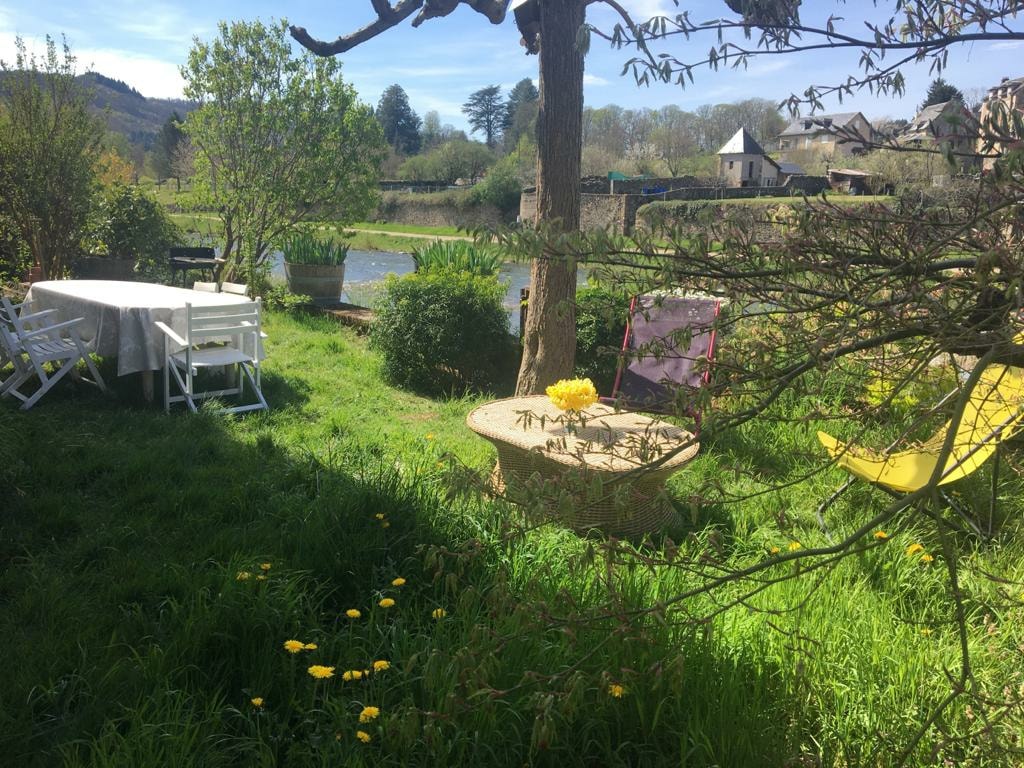 A serene garden area is highlighted by a round stone table surrounded by green grass and yellow wildflowers. Nearby, two sun loungers and a white dining table are placed under the shade of trees, with a gentle river visible in the background.