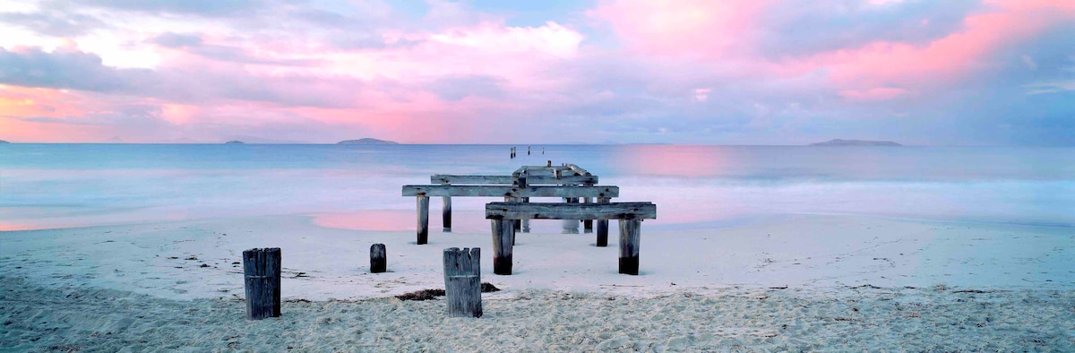 A wooden pier extends into calm waters, surrounded by soft sandy beach. The sky showcases hues of pastel pink and purple, reflecting on the water's surface. A tranquil scene invites relaxation, with gentle waves lapping against the shore.