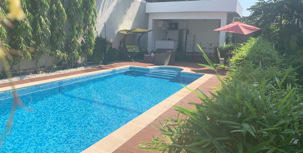A tranquil outdoor space is highlighted by a clear blue swimming pool surrounded by lush greenery. A shaded seating area features a lounge chair and an umbrella-covered table nearby, while a modern structure is visible in the background.