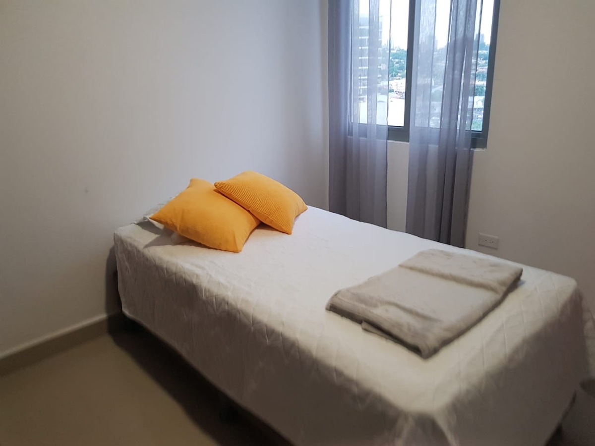A cozy bedroom is equipped with a spacious bed topped with two orange pillows. A light gray blanket is neatly folded at the foot of the bed. Natural light enters through a large window adorned with sheer curtains, enhancing the calm atmosphere.