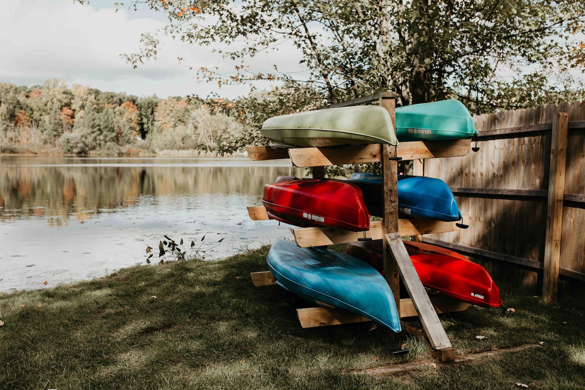 A rack holds four kayaks in varying colors of green, blue, and red, positioned beside the tranquil waters of Lake Forest. The surrounding area features lush greenery and reflections of trees in the lake, creating a serene outdoor setting.