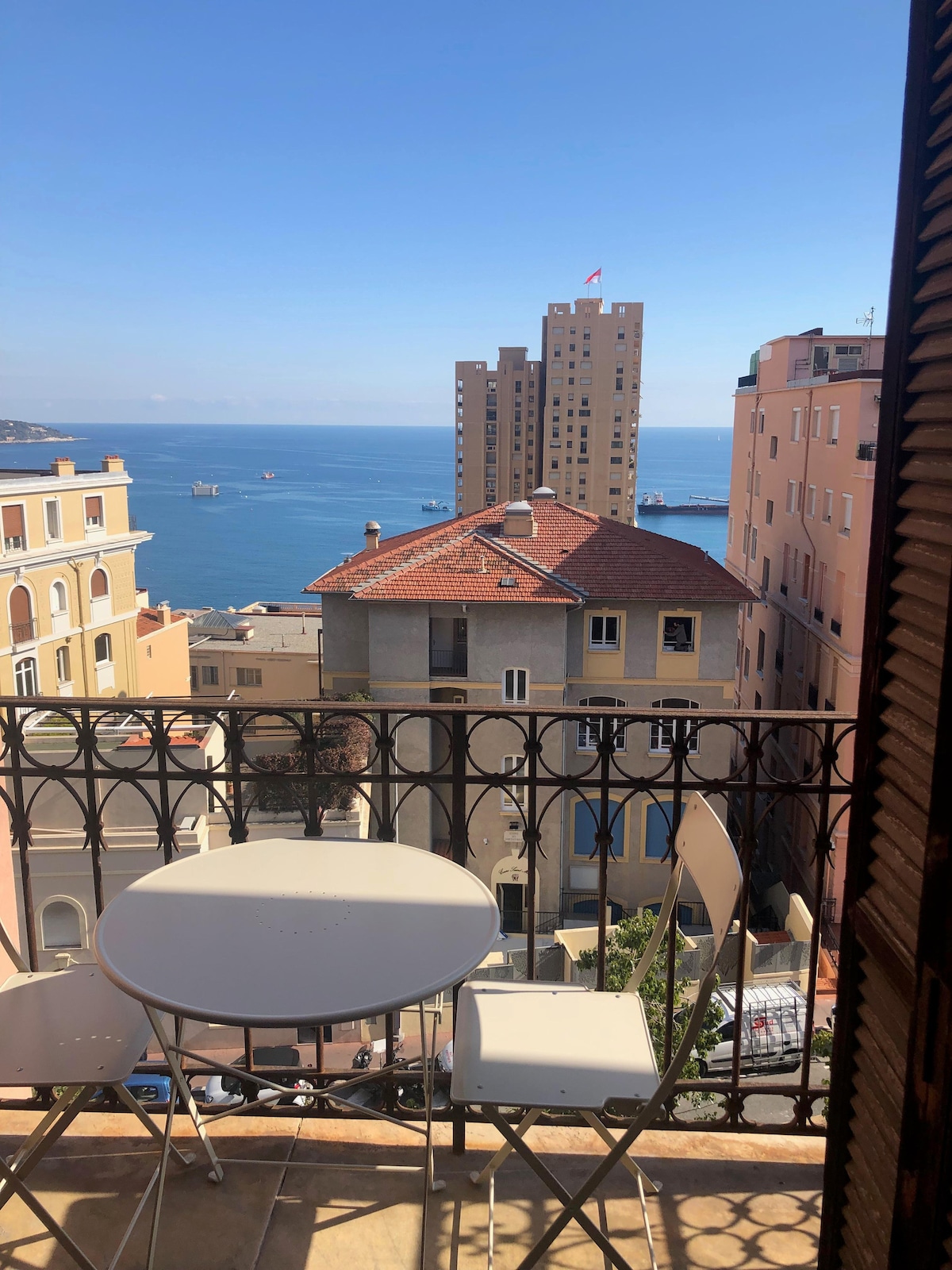 A balcony features a small round table and two chairs, overlooking a scenic view of the sea and neighboring buildings. The distant shoreline is visible under a clear blue sky, with a flag fluttering in the breeze.