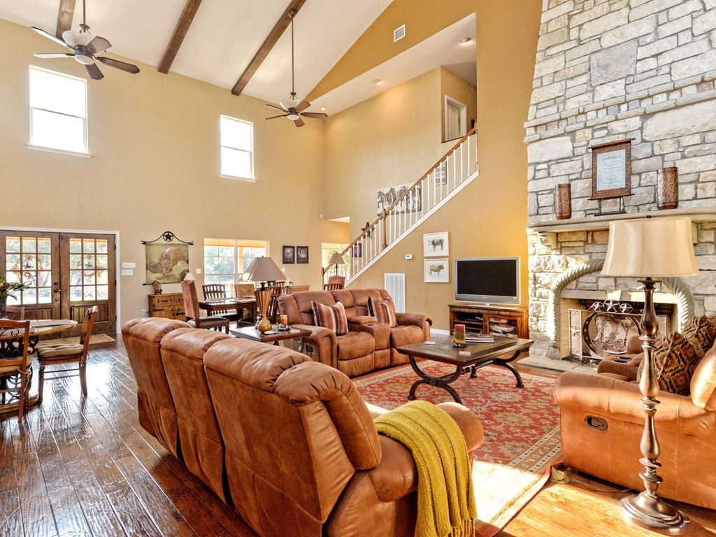 A spacious great room features high ceilings with wooden beams and a large stone fireplace. Comfortable leather seating is arranged around a coffee table, while natural light fills the space through multiple windows. A staircase and dining area can be seen in the background.
