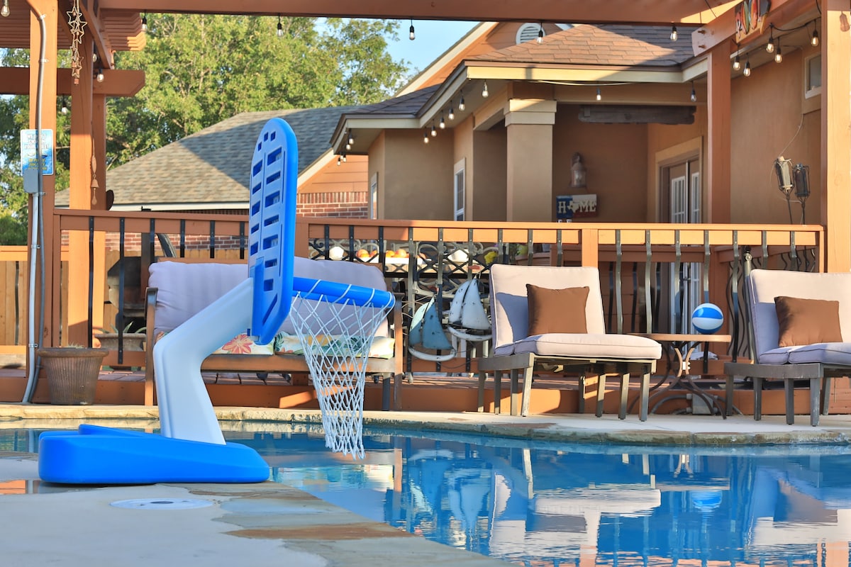 A pool area features a basketball hoop floating in the water, complemented by a spacious patio. Comfortable seating arrangements with cushions are positioned nearby, alongside decorative elements like a beach ball. Warm lighting casts a pleasant glow in the late afternoon.