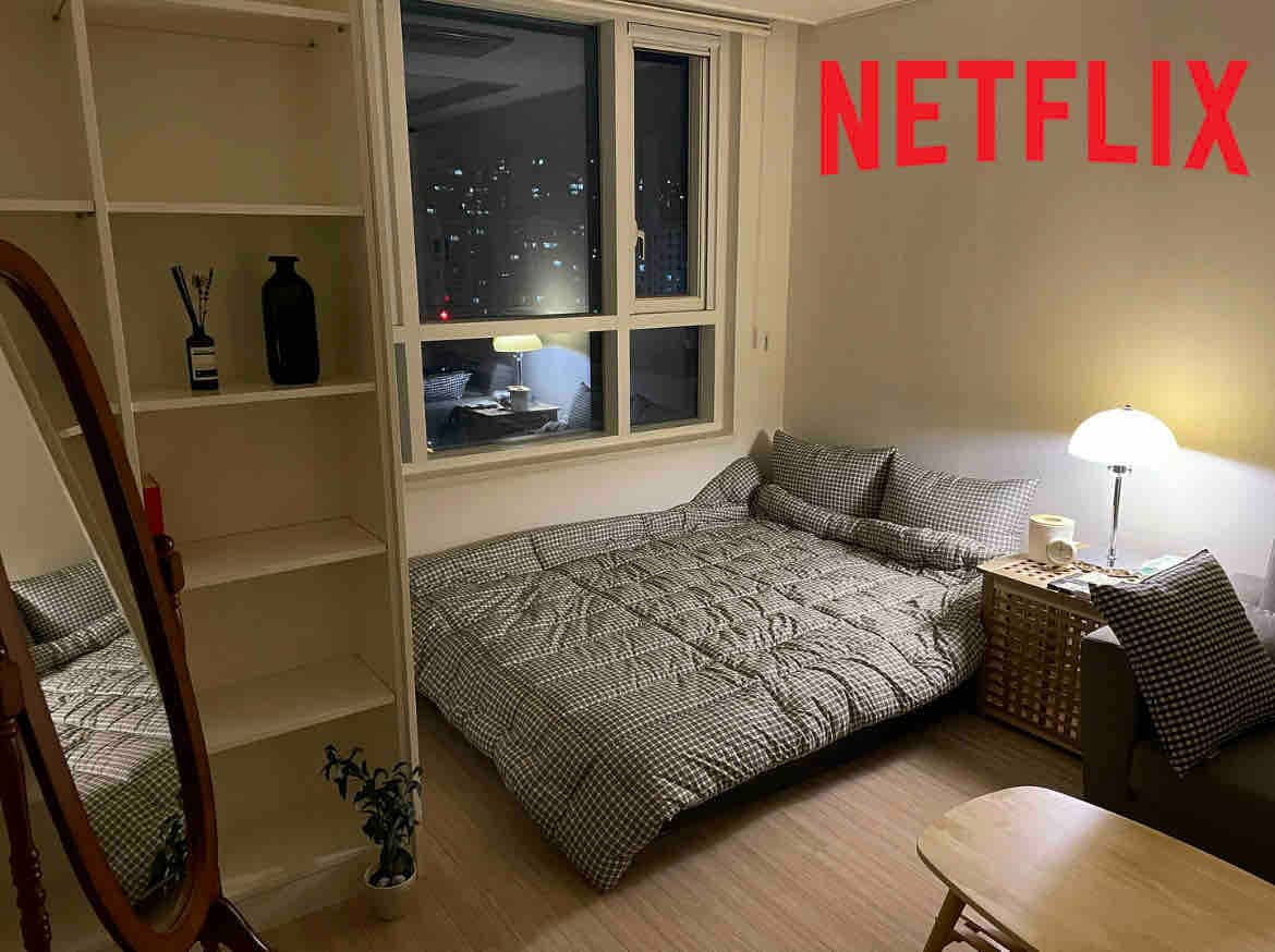 A cozy sleeping area features a spacious bed adorned with patterned bedding. A small light provides illumination next to the bed, and a mirror is positioned against the wall. A shelf displays decorative items, while a window reveals a city view in the background.