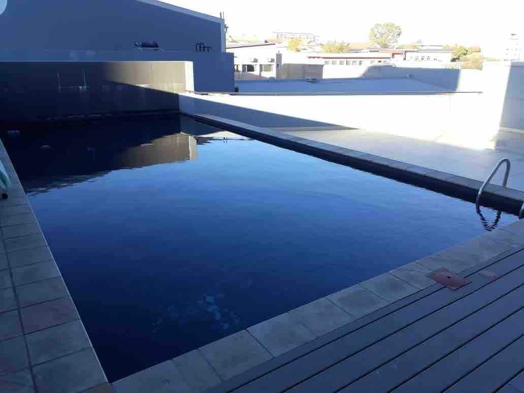 A rooftop swimming pool is presented, featuring calm blue waters surrounded by tiled decking. The pool area is set against a backdrop of the city skyline, with sunlight reflecting off the water's surface, creating a serene environment for relaxation.