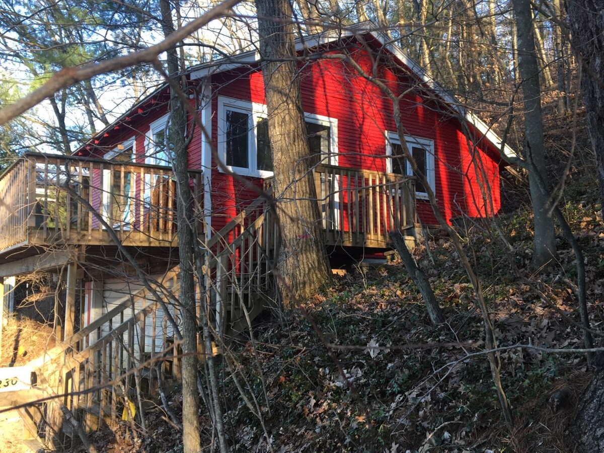 A rustic red cottage is nestled among trees on a hillside, featuring a wooden deck that extends from the main entrance. Large windows provide glimpses of the surrounding forest, while the natural landscape is enriched with fallen leaves and greenery.