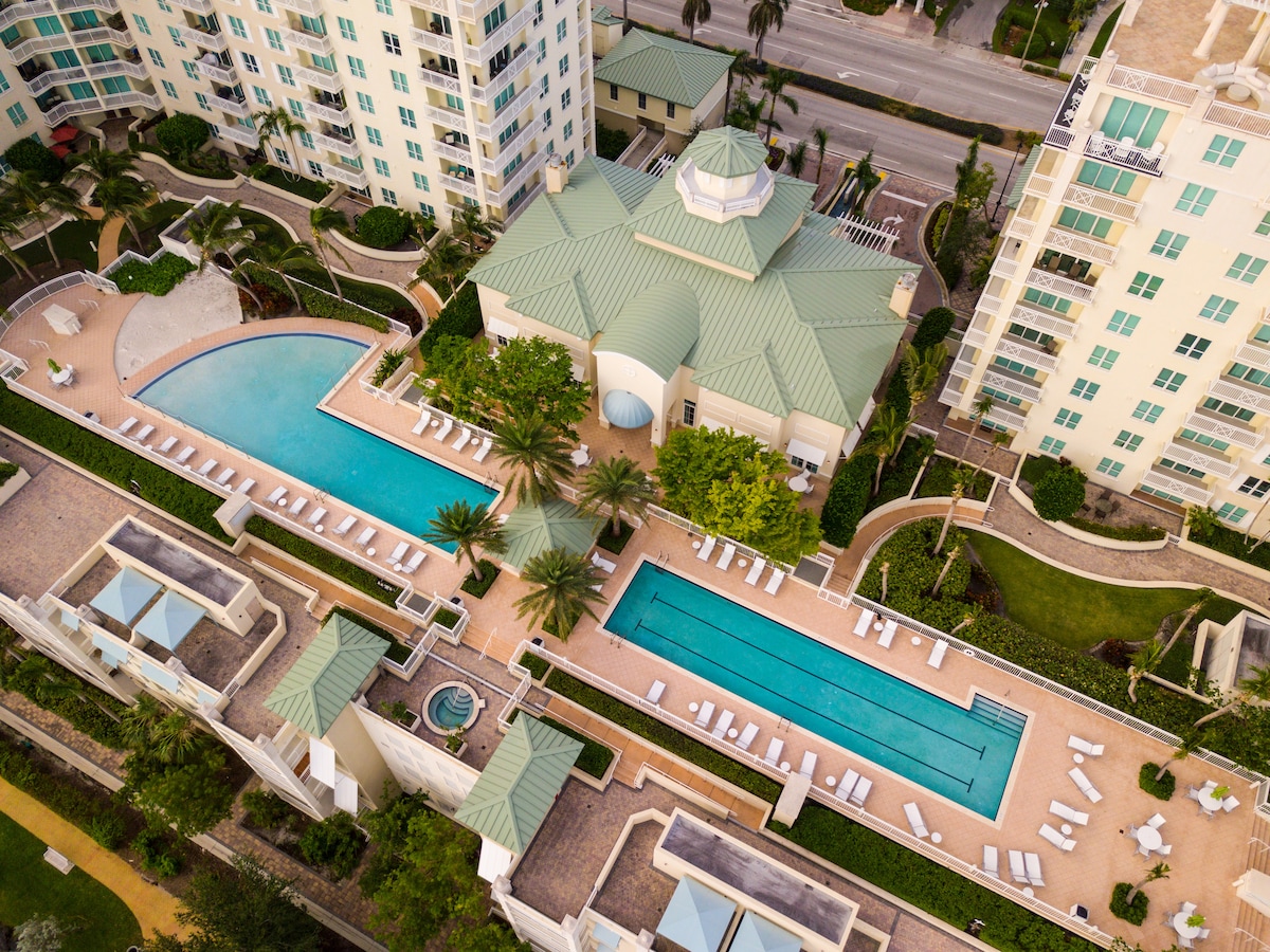 An aerial view of the community amenities showcases two large swimming pools surrounded by lounge chairs and palm trees. The clubhouse with a distinctive green roof is visible, alongside a hot tub and landscaped pathways, enhancing the resort-like atmosphere.